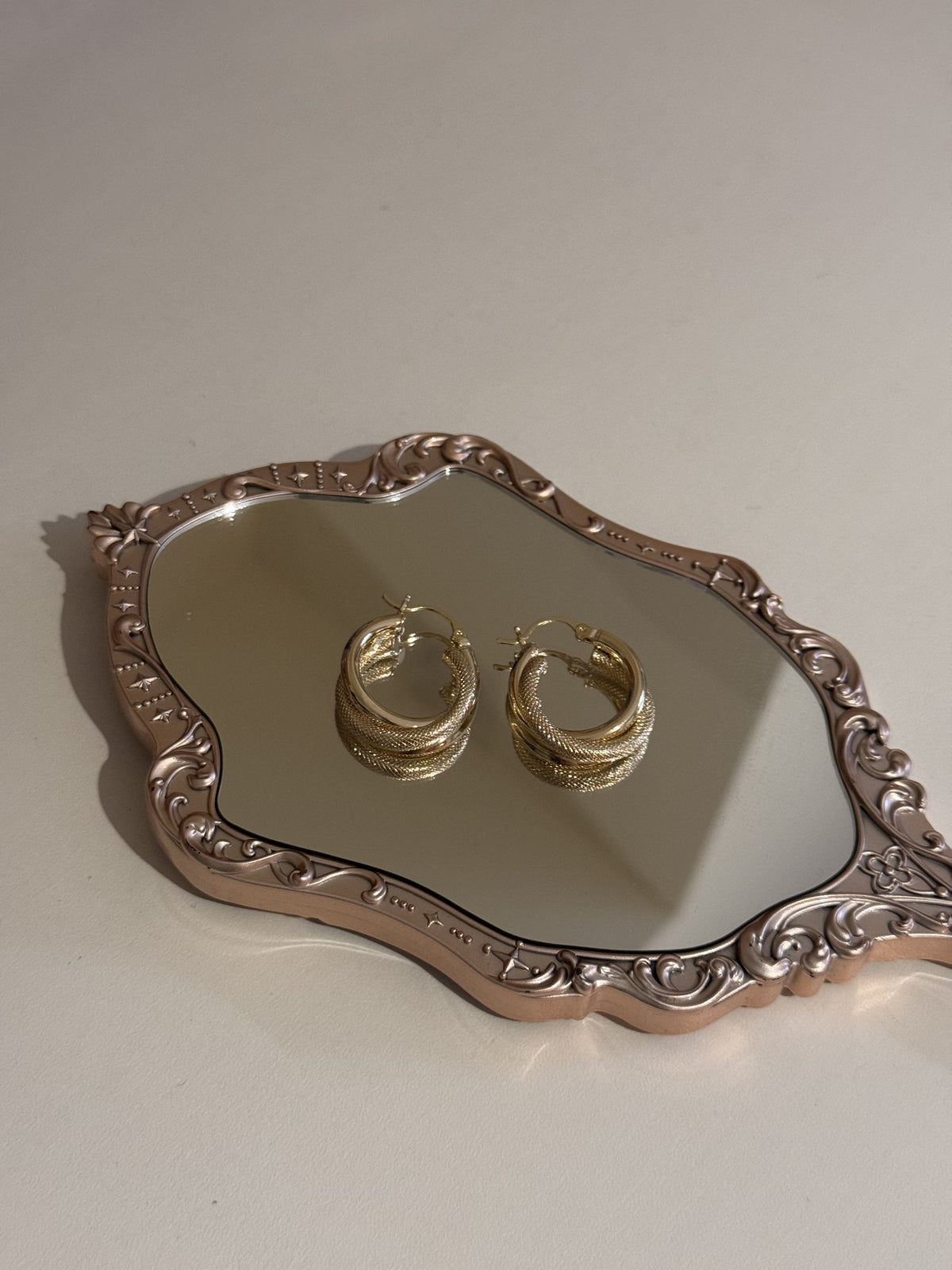 A pair of gold-tone textured hoop earrings resting on a vintage mirrored tray.