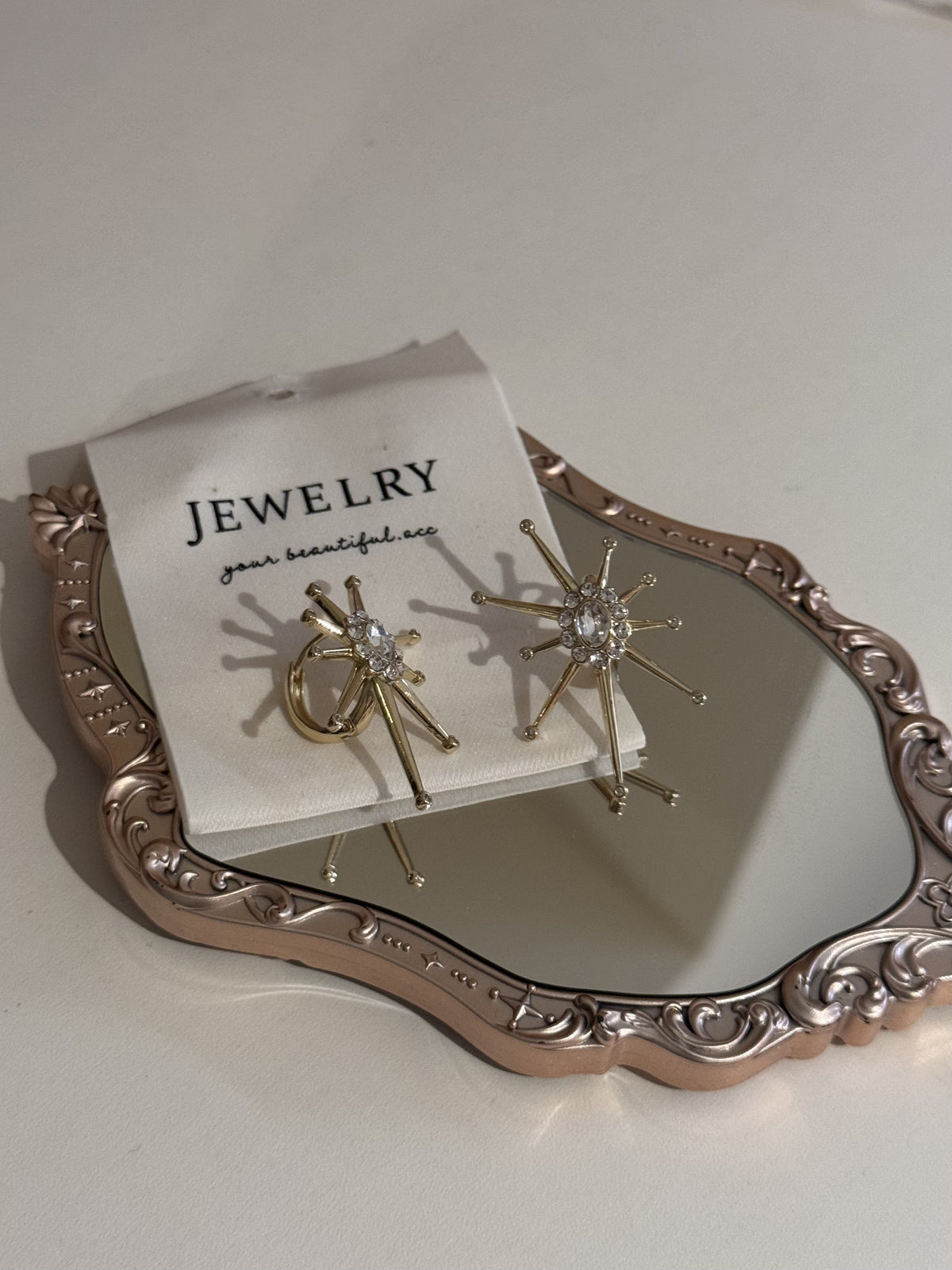 A pair of gold-tone starburst earrings with clear crystal centers displayed on a card.