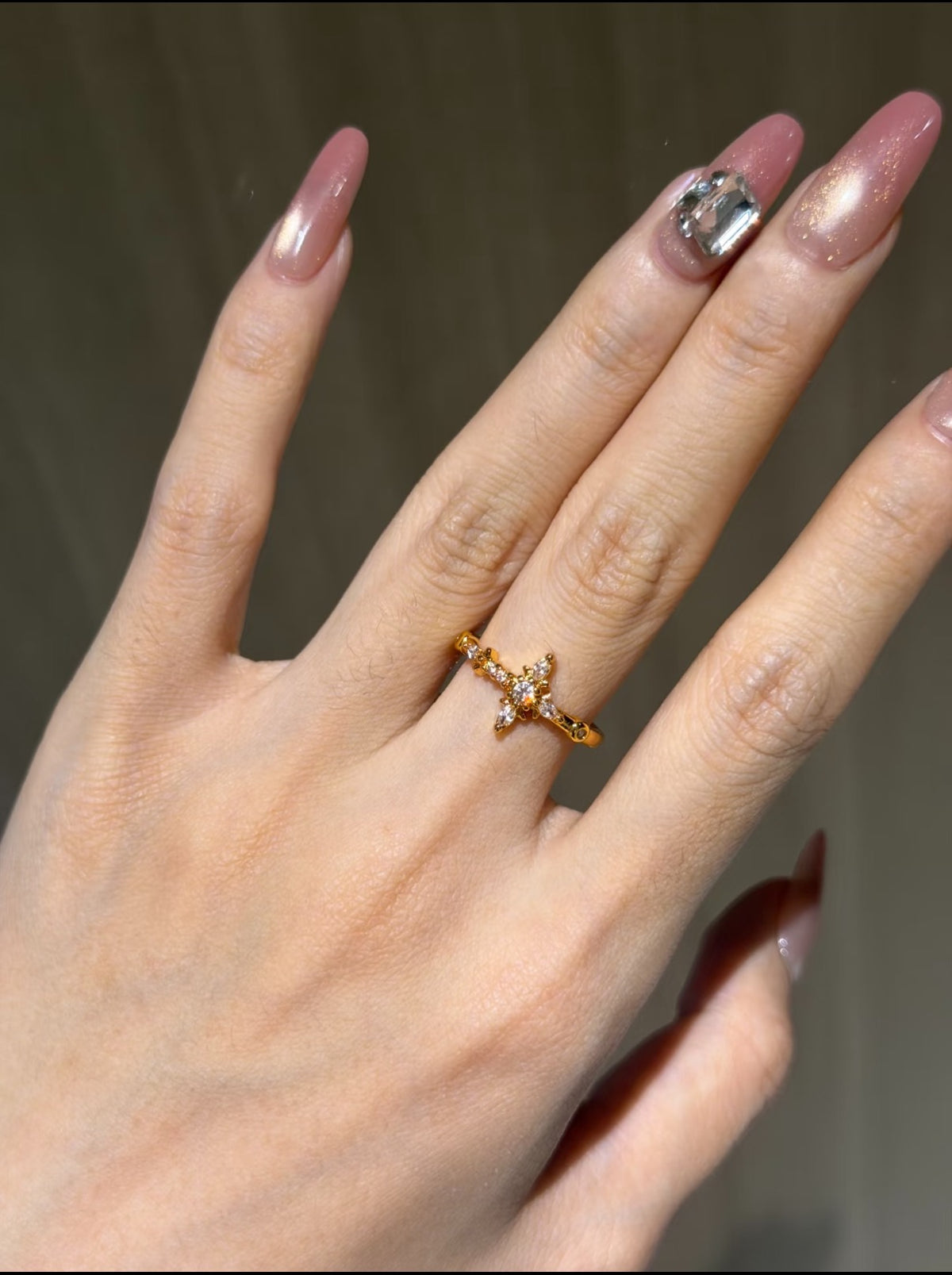 Gold cross ring with clear stones on a hand with pink manicured nails.