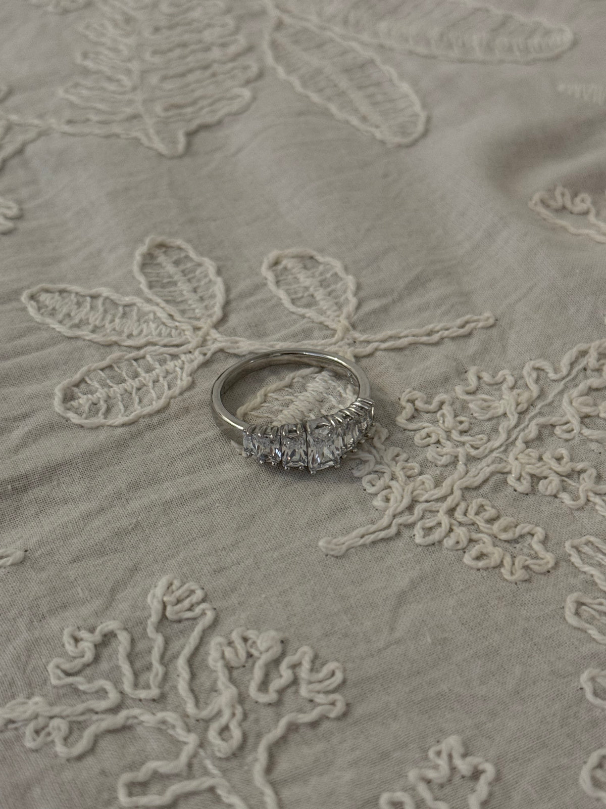 Silver-tone ring set with a row of clear rectangular crystals resting on cream embroidered linen fabric