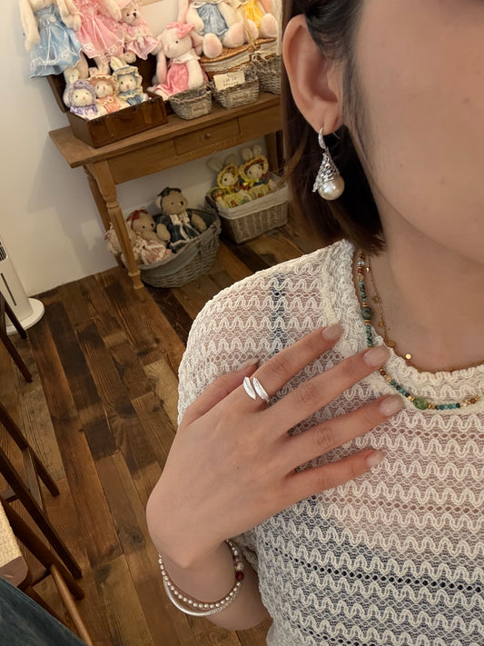 Close-up of a woman wearing pearl drop earrings, a beaded necklace, a ring, and a pearl bracelet in a doll-filled room.