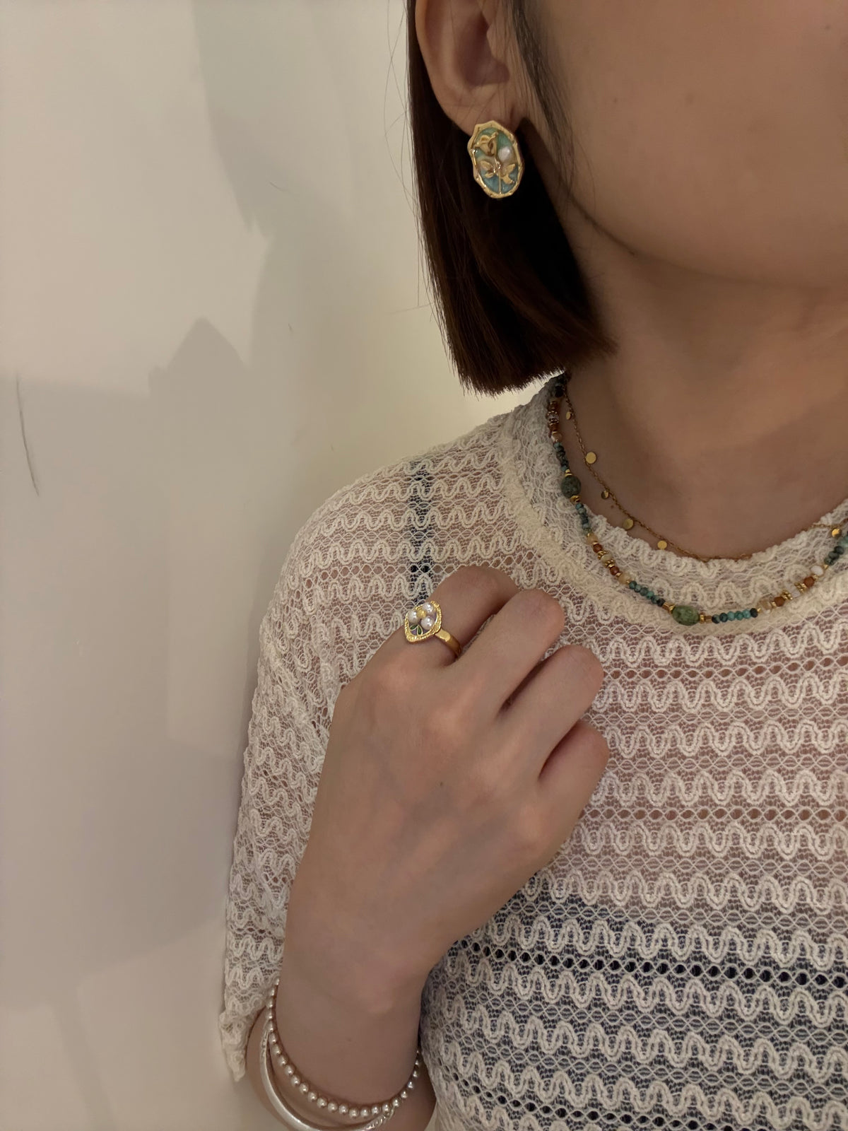 Close-up of woman wearing gold floral earrings, beaded necklace, gemstone ring, and pearl bracelet.