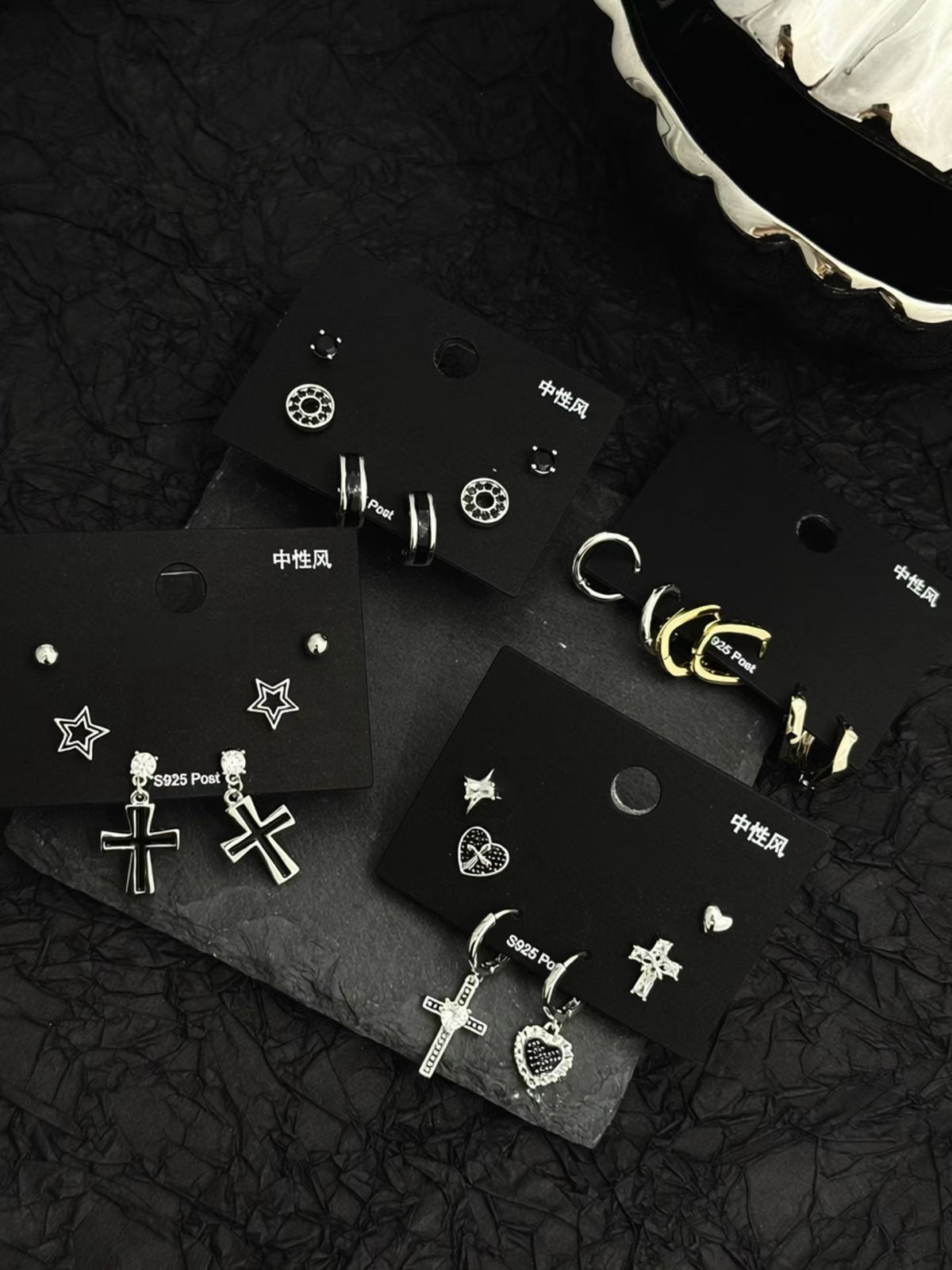 Silver earrings on black cards: crosses, stars, hearts, studs; dark background.