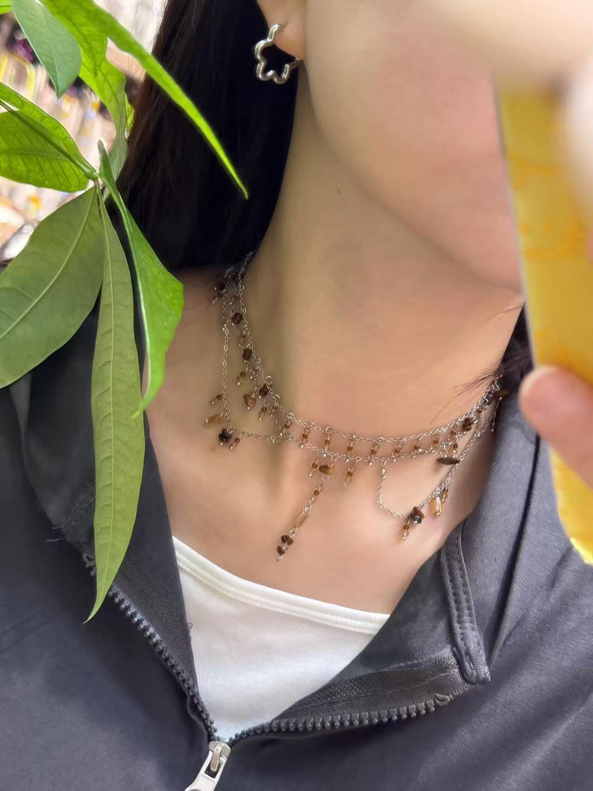 Woman wearing silver chain necklace with dangling brown beads and flower earring, leafy plant foreground