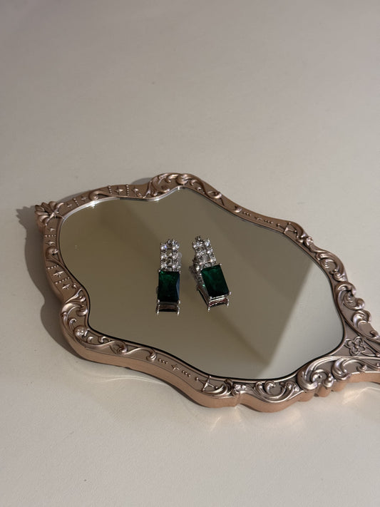 A pair of rectangular green and crystal stud earrings displayed on a decorative mirror.