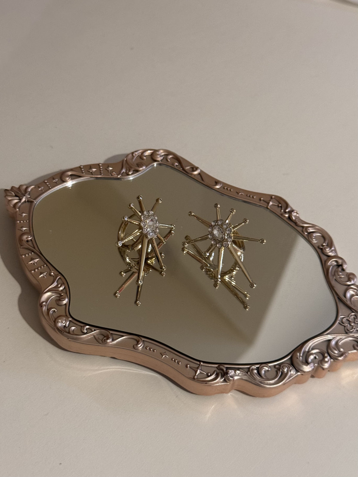 A pair of gold-tone starburst earrings with clear crystal centers resting on a mirrored tray.