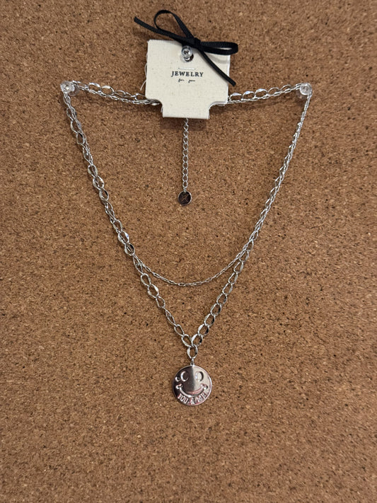 A silver-tone double chain necklace featuring a round smiley face pendant engraved with the word SMILE.