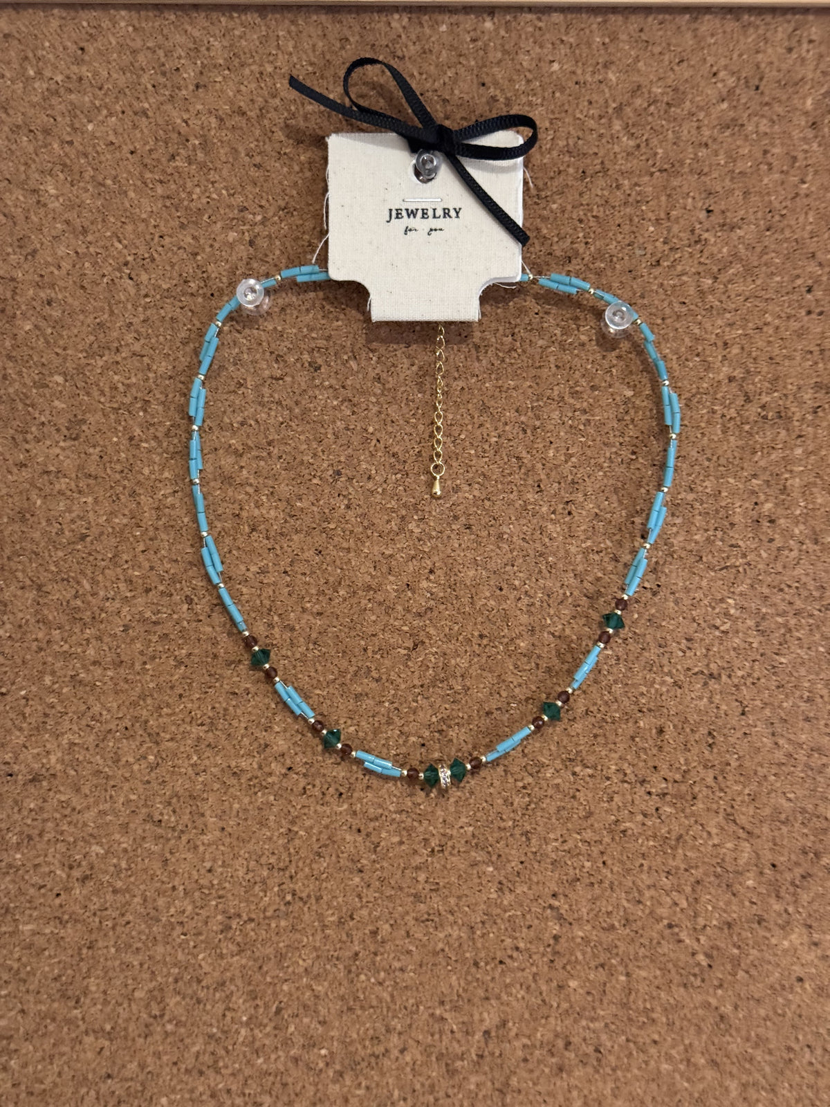 A light blue beaded necklace accented with green beads and a gold chain extender on a display card.