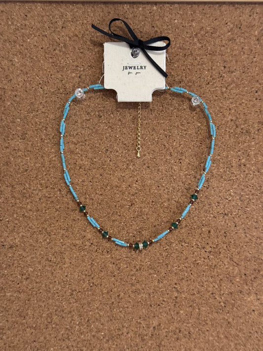 A light blue beaded necklace accented with green beads and a gold chain extender on a display card.