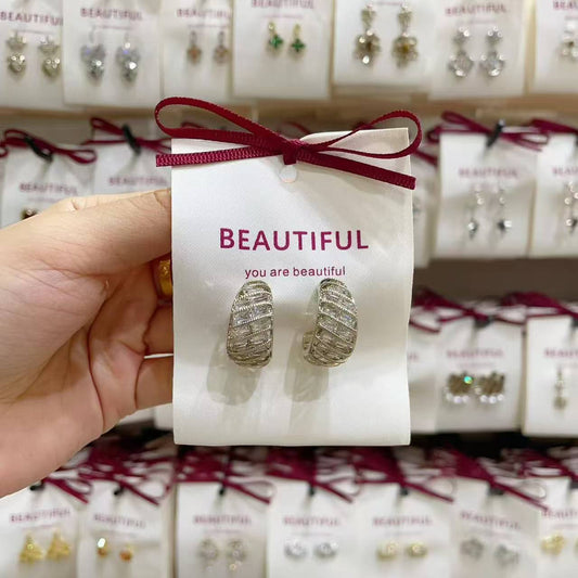 Pair of wide textured silver-toned earrings on a white BEAUTIFUL card held by hand, with jewelry display in background.